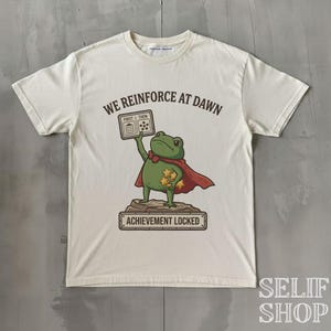 Könnte beinhalten: Ein cremefarbenes Kurzarm-T-Shirt mit einer Grafik eines grünen Frosches in einem roten Umhang, der auf einem Felsen steht. Der Frosch hält ein Schild mit der Aufschrift FIRST THEN und goldene Sterne. Text: WE REINFORCE AT DAWN und ACHIEVEMENT LOCKED.