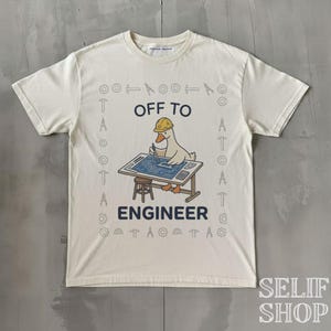 Puede incluir: Una camiseta de manga corta color hueso con un gráfico de un pato con casco amarillo, trabajando en una mesa de dibujo con planos. El texto dice OFF TO ENGINEER, rodeado por un borde de pequeños iconos de herramientas de ingeniería.