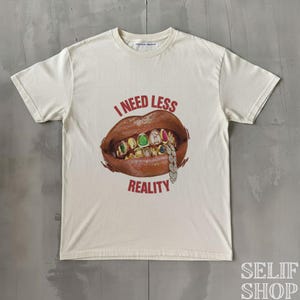 Könnte beinhalten: Cremefarbenes T-Shirt mit einem Aufdruck eines Mundes mit Goldzähnen, bunten Edelsteinen und einer Diamantkette. Der Text "I NEED LESS REALITY" ist in Rot über und unter dem Mund aufgedruckt. Das Shirt hat einen Rundhalsausschnitt und kurze Ärmel.
