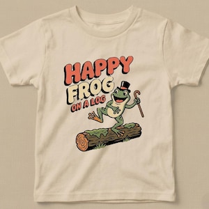 Könnte beinhalten: Cremefarbenes T-Shirt mit einem Cartoon-Frosch, der einen Zylinder trägt und einen Stock hält, auf einem Baumstamm steht. Der Text "HAPPY FROG ON A LOG" ist über dem Frosch in einer Retro-Schrift gedruckt.
