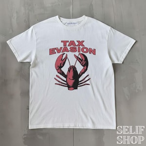 Può includere: Una t-shirt bianca a maniche corte con la grafica di un'aragosta rossa centrata sul petto. Sopra l'aragosta, il testo TAX EVASION è stampato in lettere maiuscole rosse in grassetto. La maglietta presenta un girocollo e un orlo dritto.