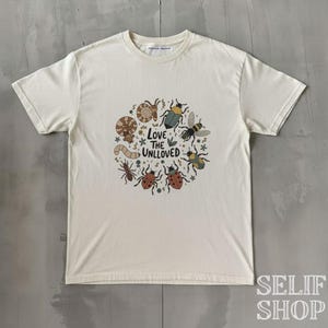 Könnte beinhalten: Cremefarbenes T-Shirt mit einem kreisförmigen Design aus verschiedenen Insekten und dem Text "Love the Unloved". Die Grafik zeigt eine Biene, Marienkäfer und andere Käfer in einem skurrilen Stil. Das Shirt hat einen Rundhalsausschnitt und kurze Ärmel.