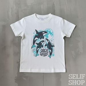 Könnte beinhalten: Weißes T-Shirt mit einer Grafik von drei Orcas und einem Vollmond. Die Orcas sind schwarz und weiß mit türkisfarbenen Details. Der Text "ORCA SPIRIT" steht darunter. Das T-Shirt hat einen Rundhalsausschnitt und kurze Ärmel.