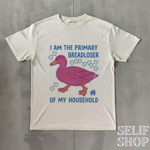 Könnte beinhalten: Ein cremefarbenes Kurzarm-T-Shirt mit einer Grafik einer rosa Ente, umgeben von kleinen Brotscheiben-Symbolen. Der Text lautet I AM THE PRIMARY BREADLOSER OF MY HOUSEHOLD in blauen Buchstaben, ergänzt durch ein kleines blaues Haussymbol.