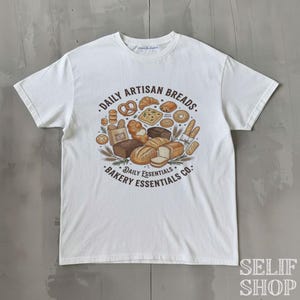 Könnte beinhalten: Weißes T-Shirt mit einer runden Grafik verschiedener Backwaren, darunter Brotlaibe, Brezeln und Croissants. Der Text lautet "Daily Artisan Breads. Bakery Essentials Co."