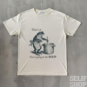 Könnte beinhalten: Cremefarbenes T-Shirt mit einer Illustration eines Frosches, der in einem Topf rührt. Der Text "That's it. You're going in the SOUP" ist oberhalb und unterhalb der Frosch-Illustration gedruckt. Rundhalsausschnitt und kurze Ärmel.