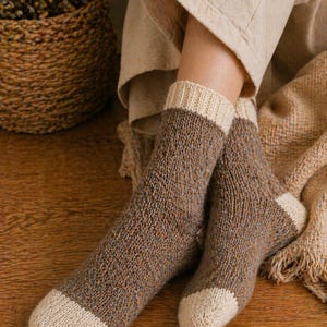 May include: A pair of brown and beige knit socks. The socks have a textured pattern with beige toes, heels, and cuffs. The socks are worn by a person sitting on a wooden surface.