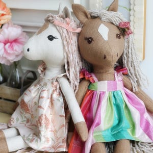 May include: Two plush horse dolls with yarn manes. The left doll is cream-colored with a floral dress and pink ribbon. The right doll is brown with a white forehead patch, wearing a dress with vertical stripes in pink, green, and white.