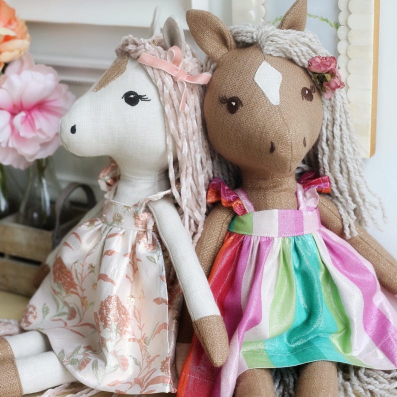 May include: Two plush horse dolls with yarn manes. The left doll is cream-colored with a floral dress and pink ribbon. The right doll is brown with a white forehead patch, wearing a dress with vertical stripes in pink, green, and white.