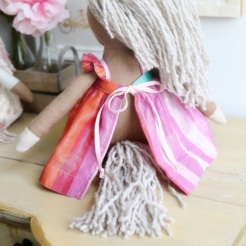 May include: A fabric doll with tan skin and long, textured off-white yarn hair. The doll wears a sleeveless dress featuring vertical stripes in shades of orange, red, and pink, with a light pink ribbon tied in a bow at the center of the back.