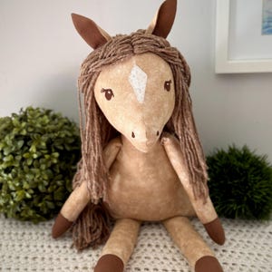 May include: A plush horse toy made of tan fabric with a mottled texture. It features a white diamond-shaped patch on its forehead, dark brown ears, and dark brown hooves. The mane is crafted from thick, brown yarn strands that hang down.