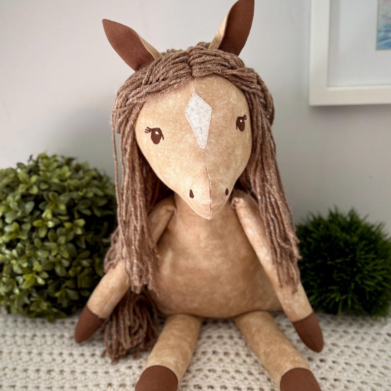 May include: A plush horse toy made of tan fabric with a mottled texture. It features a white diamond-shaped patch on its forehead, dark brown ears, and dark brown hooves. The mane is crafted from thick, brown yarn strands that hang down.