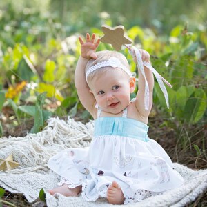 May include: A baby girl wearing a white dress with a blue bodice and a floral headband holds a wooden star wand with a white ribbon. She is sitting on a white blanket in a grassy area.