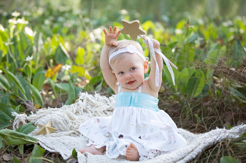 May include: A baby girl wearing a white dress with a blue bodice and a floral headband holds a wooden star wand with a white ribbon. She is sitting on a white blanket in a grassy area.