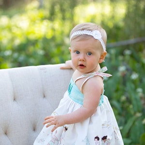May include: A baby girl wearing a white headband with a flower, a light blue and white dress with a floral pattern and a lace trim, and a white bow in her hair. She is sitting on a white chair with a tufted back.