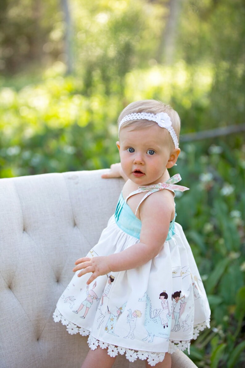 May include: A baby girl wearing a white headband with a flower, a light blue and white dress with a floral pattern and a lace trim, and a white bow in her hair. She is sitting on a white chair with a tufted back.