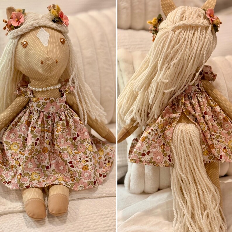 May include: A fabric horse doll with a cream-colored body, white yarn mane, and a floral print dress in shades of pink, yellow, and green. The doll features a white forehead patch, a pearl-style necklace, and small fabric flowers in its hair.
