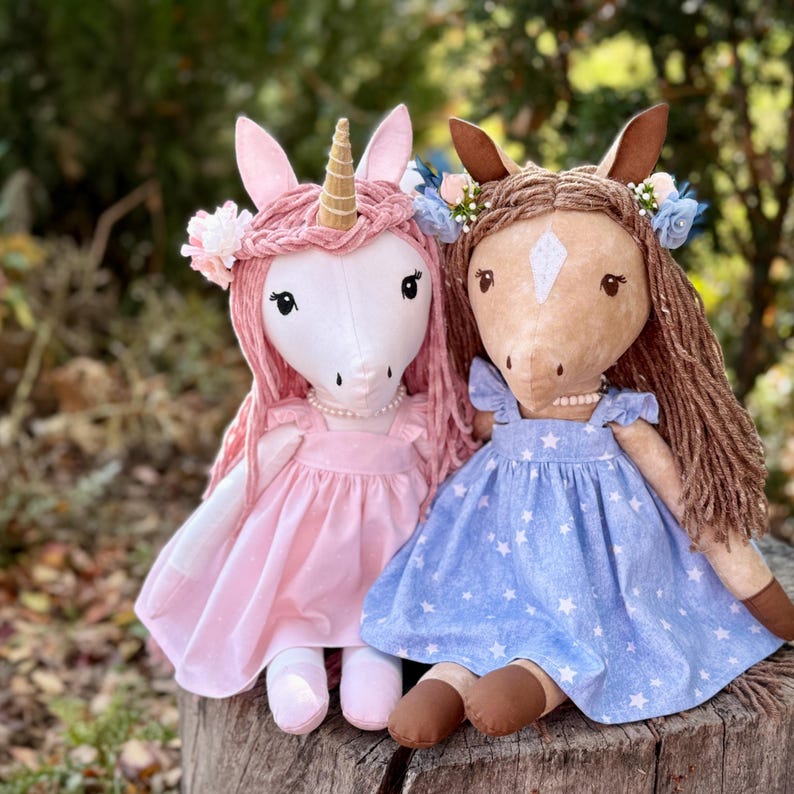 May include: Two plush dolls, one a white unicorn with a gold horn and pink yarn hair, the other a tan horse with brown yarn hair. Both wear pearl necklaces and floral headbands. The unicorn wears a pink dress, the horse a blue star-print dress.