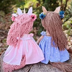 May include: Two handmade dolls with yarn hair. The left doll has pink yarn hair, a gold horn, and a pink dress with a white ribbon. The right doll has brown yarn hair and a blue dress with a white star pattern. Both dolls feature floral hair accessories.