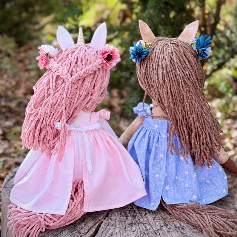 May include: Two handmade dolls with yarn hair. The left doll has pink yarn hair, a gold horn, and a pink dress with a white ribbon. The right doll has brown yarn hair and a blue dress with a white star pattern. Both dolls feature floral hair accessories.
