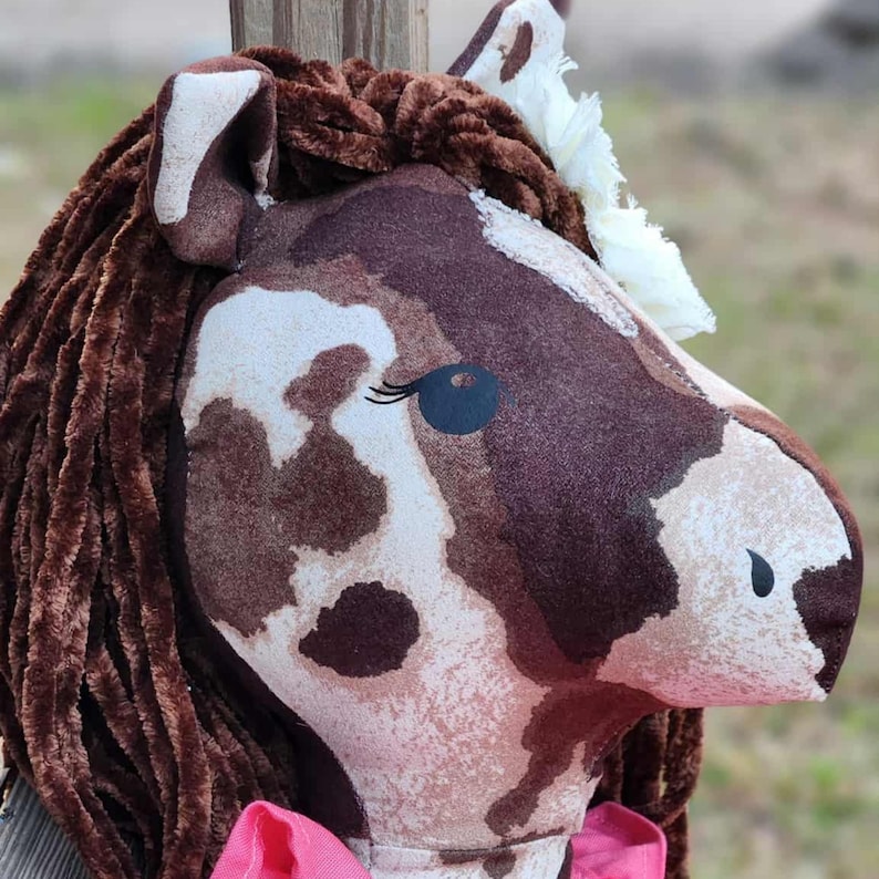 May include: A plush horse head toy featuring a brown and white mottled pattern, dark brown textured yarn mane, white fabric forelock, black eye detail with eyelashes, and a bright pink fabric accent at the base of the neck.