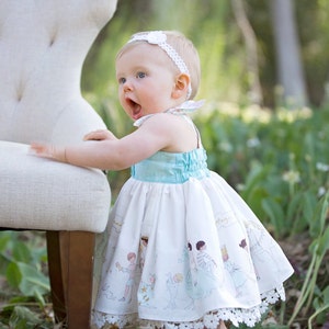 May include: A baby girl wearing a white dress with a blue bodice and a white headband. The dress has a print of children playing. The baby is standing next to a white chair.
