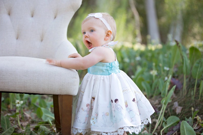 May include: A baby girl wearing a white dress with a blue bodice and a white headband. The dress has a print of children playing. The baby is standing next to a white chair.