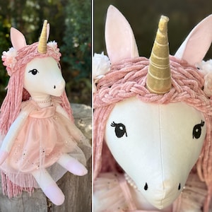 May include: A plush unicorn doll with white fabric skin, a gold metallic horn, and long pink yarn hair. The doll wears a braided pink yarn headband with small white flowers, a pearl necklace, and a pink dress with a sheer, glittery tulle overlay.