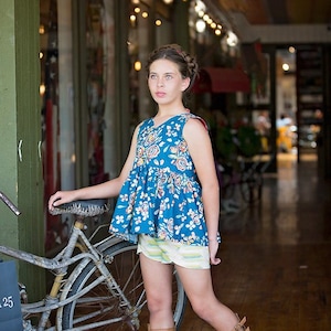 Könnte beinhalten: Eine junge Person trägt ein blaues und weißes, ärmelloses Oberteil mit Blumenmuster und einen weißen und gelben, gestreiften Rock, braune Gladiatorsandalen und eine geflochtene Frisur. Sie steht neben einem Fahrrad mit braunem Sattel und einem schwarzen Korb.