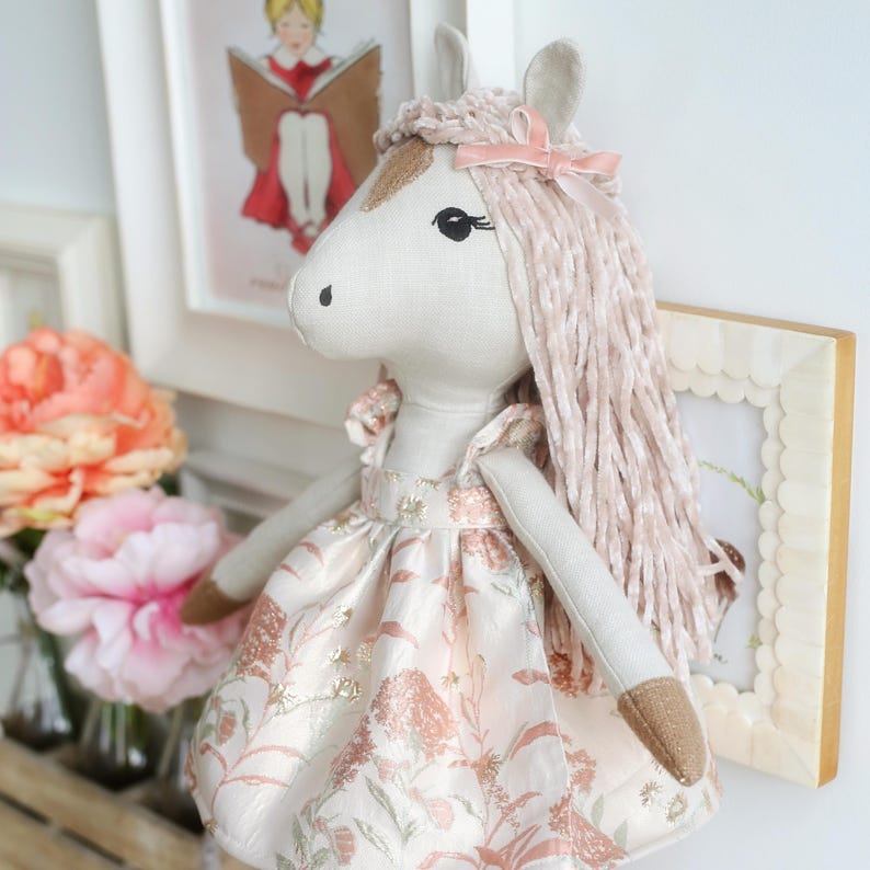May include: A plush horse doll with a light beige fabric body, long textured pale pink yarn hair, and a small pink ribbon bow. The doll wears a dress featuring a floral pattern in shades of pink, white, and metallic gold with ruffled sleeves.