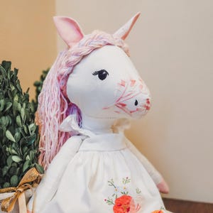 May include: A plush unicorn doll with white fabric skin, pink ears, and long, braided pastel yarn hair. The face features embroidered black eyes and pink floral stitching on the snout. It wears a white dress with embroidered red and green flowers.