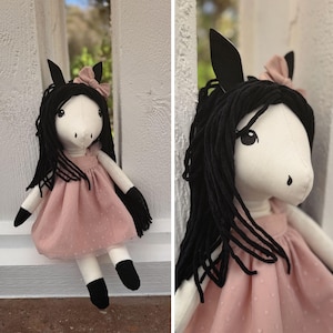 May include: A plush horse doll with a white fabric body, black yarn hair, and black ears. The doll wears a dusty pink dress with a subtle dotted texture and a matching pink bow on its head. The doll has black fabric hooves on its arms and legs.