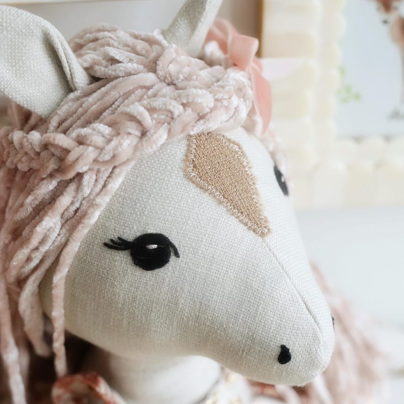 May include: A plush horse toy made of light beige fabric with a soft, textured, pale pink velvet mane styled in a braid. The horse features embroidered black eyes with eyelashes, a tan diamond-shaped patch on the forehead, and a small pink ribbon.