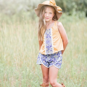 Peut inclure: Une jeune fille portant un haut à imprimé floral jaune et violet avec un chapeau brun et des sandales gladiateur brunes. Elle se tient dans un champ d'herbe haute.