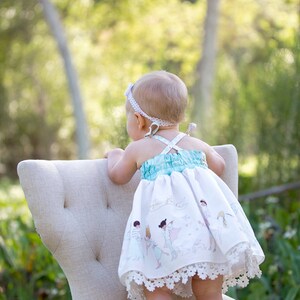 May include: A baby girl wearing a white dress with a blue bodice and a white lace trim stands on a white chair. The dress has a print of children playing. The baby is looking away from the camera.