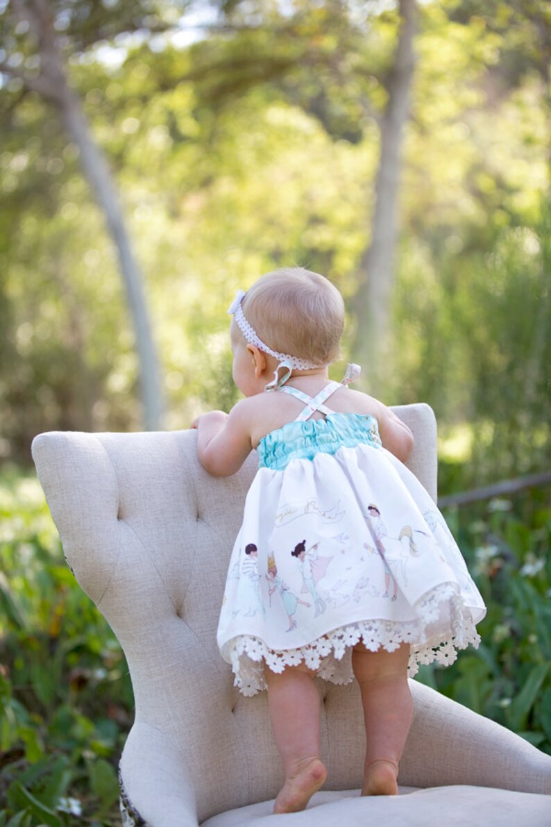May include: A baby girl wearing a white dress with a blue bodice and a white lace trim stands on a white chair. The dress has a print of children playing. The baby is looking away from the camera.