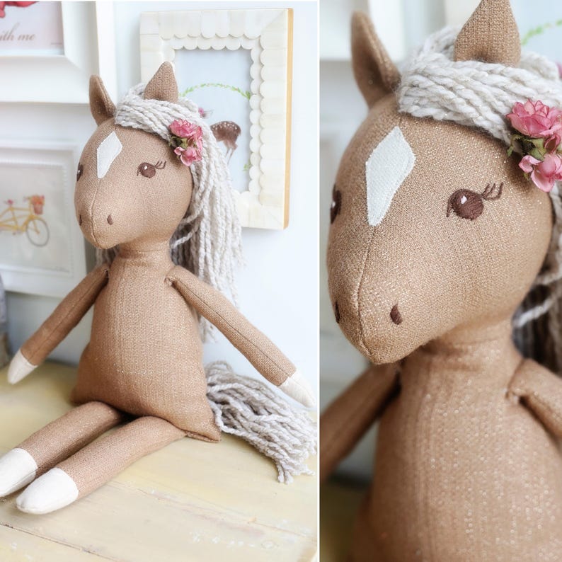 May include: A tan fabric horse doll with a white diamond-shaped patch on its forehead, white fabric hooves, and a long, thick, cream-colored yarn mane and tail. The horse features embroidered brown eyes with eyelashes and small pink fabric flowers.