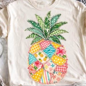 Puede incluir: Camiseta color crema con un diseño de piña de patchwork colorido. La piña tiene hojas verdes y un cuerpo compuesto por varias telas estampadas, incluyendo cuadros, lunares e impresiones florales. La camiseta tiene mangas cortas.