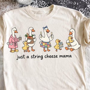 May include: Cream-colored t-shirt featuring a whimsical illustration of geese and ducklings. The geese are depicted in various outfits, holding items like coffee, a basket of food, and string cheese. The text "just a string cheese mama" is printed below the illustration.