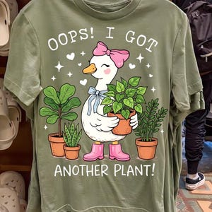 Puede incluir: Camiseta verde salvia con un dibujo de un ganso de dibujos animados con un lazo y botas rosas, sosteniendo una planta en maceta. La camiseta tiene el texto "OOPS! I GOT ANOTHER PLANT!" con plantas en maceta y estrellas adicionales.