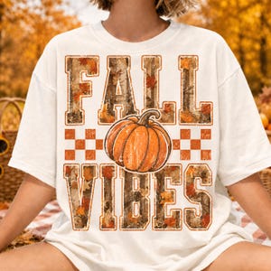 May include: An off-white short-sleeve t-shirt featuring the text FALL VIBES in large block letters. The letters have a camouflage and autumn leaf pattern. A central orange pumpkin graphic sits between the words, with a checkered pattern.