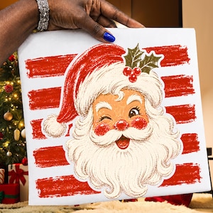 May include: A square white canvas featuring a graphic illustration of a winking Santa Claus with a red hat, rosy cheeks, and a white beard. The background consists of horizontal red brushstroke stripes, and a sprig of holly is placed on the hat.