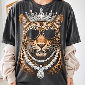 May include: A charcoal gray short-sleeve t-shirt featuring a large graphic print of a leopard wearing black sunglasses, a jeweled crown, and a layered diamond necklace. The leopard has brown fur with black spots and long white whiskers.