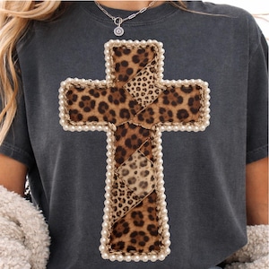 Könnte beinhalten: Dunkelgraues T-Shirt mit einem großen Kreuzdesign. Das Kreuz besteht aus Leopardenmusterstoff mit einem Kunstperlenrand. Das Kreuz ist mit einem goldenen Faden genäht. Das Shirt ist ein lässiger Stil.