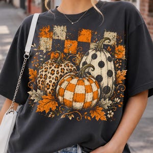May include: A charcoal gray short-sleeve t-shirt featuring a graphic print of three pumpkins with different patterns including leopard print, orange and white checkered, and black polka dots, surrounded by orange autumn leaves.