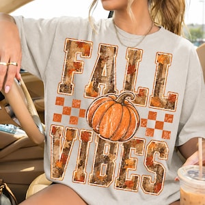 May include: A light gray short-sleeve t-shirt featuring the text FALL VIBES in large block letters filled with a camouflage and autumn leaf pattern. A central orange pumpkin graphic sits between the words, accented by a checkered pattern.