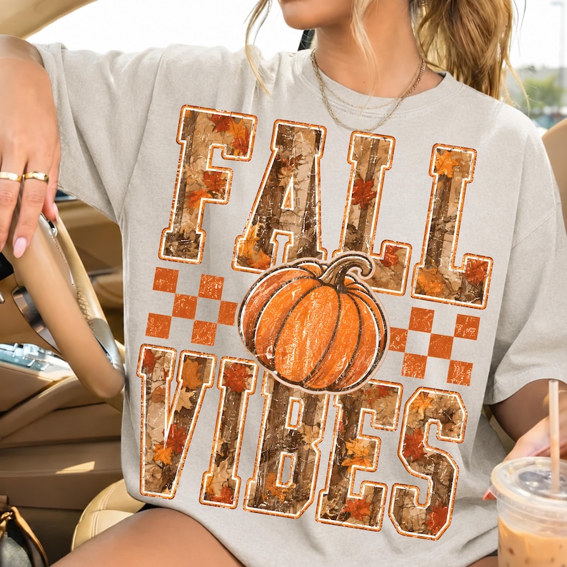 May include: A light gray short-sleeve t-shirt featuring the text FALL VIBES in large block letters filled with a camouflage and autumn leaf pattern. A central orange pumpkin graphic sits between the words, accented by a checkered pattern.