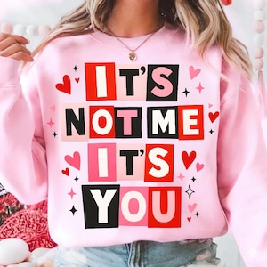 Puede incluir: Sudadera rosa claro con el texto "IT'S NOT ME IT'S YOU" en un diseño de letras en bloque. El diseño incluye bloques rojos, negros y rosas, corazones y estrellas. Una prenda informal con temática de San Valentín.
