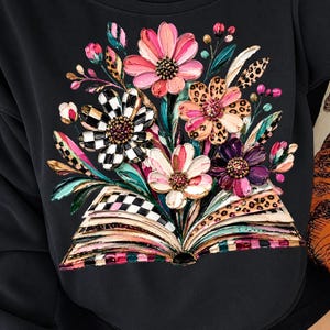 Könnte beinhalten: Ein schwarzes Sweatshirt mit einem Grafikdruck eines aufgeschlagenen Buches mit aufgefächerten Seiten. Ein lebendiger Blumenstrauß entspringt dem Buch, mit rosa, weißen, violetten und schwarz-weiß karierten Blütenblättern sowie Blättern mit Leopardenmuster.