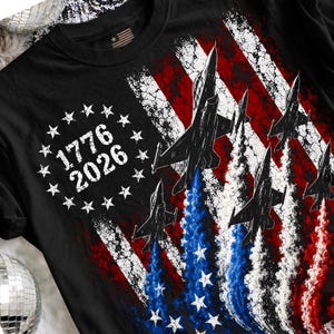 Pu&ograve; includere: T-shirt nera con un design a bandiera americana invecchiata, jet e gli anni 1776 e 2026. La bandiera utilizza i colori rosso, bianco e blu, con jet che salgono da scie di fumo blu e bianco.